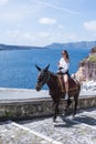 Young beautiful girl in a white blouse and shorts on a brown horse against the blue sky and the island Royalty Free Stock Photo