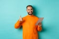 Satisfied adult man smiling, using digital tablet and showing thumb-up, approve and agree, standing against turquoise