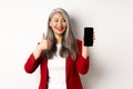 Satisfied asian elderly businesswoman showing blank smartphone screen and thumb-up, praising online promotion or company