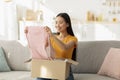 Satisfied asian woman sitting on sofa and opening carton box, checking her delivery, holding new clothes and smiling