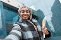 Satisfied attractive islamic afro american woman student in hijab and coat with books looks at gadget webcam