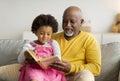 Satisfied black little kid and elderly man reading book with fairy tales on sofa in living room interior Royalty Free Stock Photo