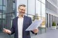 Satisfied businessman in suit holding business papers, standing in front of office on street, successful negotiation Royalty Free Stock Photo