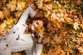 Satisfied caucasian millennial red-haired lady in raincoat lies on ground in park, closes eye with yellow leaf