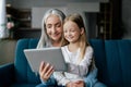 Satisfied caucasian small girl and old lady watch video, enjoy online game on tablet, have call on sofa Royalty Free Stock Photo