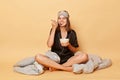 Satisfied cheerful positive young girl sitting floor with blanket wearing black pajama and sleep eye mask holding plate in hands Royalty Free Stock Photo