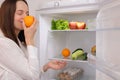 Satisfied delighted Caucasian brown haired woman standing near open refrigerator full of fruits and vegetables taking fresh orange Royalty Free Stock Photo