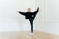 Satisfied female ballerina balances on one leg, stretches in studio, practices yoga, demonstrates good flexibility, dressed in