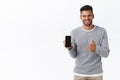 Satisfied handsome bearded male model in grey sweater, show thumb-up and smiling in approval, holding mobile phone
