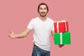 Satisfied handsome bearded young hipster man in white shirt and casual hat standing, holding two present box with raised arms,