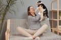 Satisfied happy smiling Caucasian woman wearing striped shirt sitting on the sofa raising her black and white puppy having fun Royalty Free Stock Photo
