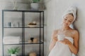 Satisfied healthy European woman poses near wall in bathroom, wrapped in white soft towels, holds mug of tea, being relaxed after