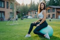 Satisfied healthy woman with curly dark hair sits on fitness ball, takes rest after yoga exercising, poses outdoor against private Royalty Free Stock Photo