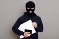 Satisfied man robber steals laptop beech and phone, being dressed in robber mask and black turtleneck, looks at camera firmly