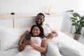 Satisfied millennial african american male and female on bed in bedroom interior look at smartphone, watch video