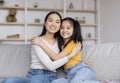 Satisfied pretty millennial chinese woman and teenage girl hugging, sitting on sofa in living room