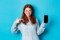 Satisfied redhead girl pointing at phone screen, showing smartphone app or online promo and smiling, standing in sweater