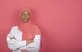 Satisfied religious female model has arms folded, looks aside, wears pink headscarf standing on pink background, copy space for