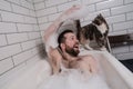 Satisfied, smiling man bathes in a bath with lush foam when his beloved cat approached him and looks with interest.