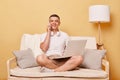 Satisfied smilng man wearing white T-shirt and shorts sitting on sofa against beige background remote job working on computer