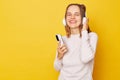 Satisfied teen girl wearing jumper and headphones posing isolated over yellow background enjoying listening music, happy to renew Royalty Free Stock Photo