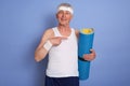 Satisfied white haired male with yoga mat posing isolated over blue background, pointing with index finger aside, wearing