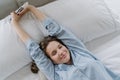 Satisfied woman stretches in bed, dressed in nightclothes, closes eyes with pleasure, holds cell phone, listens music in earphones Royalty Free Stock Photo