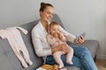Satisfied woman wearing white shirt and jeans sitting on sofa with her baby daughter, holding cell phone in hands, having online Royalty Free Stock Photo