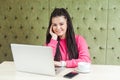 Satisfied young girl freelancer with black dreadlocks hairstyle in pink blouse sitting in cafe, working on laptop with toothy Royalty Free Stock Photo