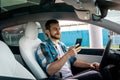 Satisfied young man sitting in the car while holding mobile phone and looking at screen. Transport, vehicle, trip and Royalty Free Stock Photo