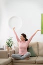 Satisfied young woman sitting on couch at home stretching out with raised hands after day sleeping