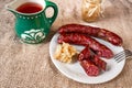 Sausage on white plate, grated horseradish with apple in small bowl, fork, green pitcher on jute tablecloth
