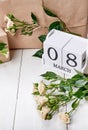 Save the date March 8 on wooden cube calendar, gift boxes wrapped in craft and fresh pastel roses on white table. Holiday concept