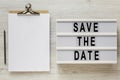 `Save the date` words on a lightbox, clipboard with blank sheet of paper on a white wooden surface, top view. Overhead, from abo Royalty Free Stock Photo