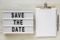 `Save the date` words on a modern board, clipboard with blank sheet of paper on a white wooden surface, top view. Overhead, from