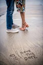 save the date written on the beach with prewedding couple standing on the background making love