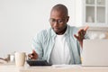 Savings and finances concept. Shocked black mature man calculating bills, using calculator, sitting in kitchen Royalty Free Stock Photo