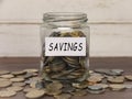 Savings label on coin jar on top of wooden desk with asorted coins background.