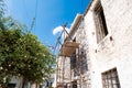 Scaffolding against a traditional house in Greece surrounded by green bushes with flowers against blue sky.