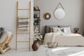 Scandi open space bedroom interior with bed with knit blanket and many pillows, rack with books and decor, carpet on the