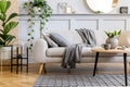 Scandinavian concept of living room interior with design sofa, coffee table, plant in pot, flowers, carpet, plaid, pillow, shelf.