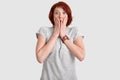 Scared emotional redhead woman covers mouth with both hands, has eyes full of fear, dressed in grey t shirt, isolated over white