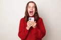 Scared frightened Caucasian woman wearing warm red sweater using phone isolated over gray background looking at camera screaming Royalty Free Stock Photo