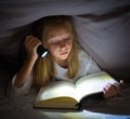 Scared little girl reading scary book under bed cover holding a flashlight late at night Royalty Free Stock Photo