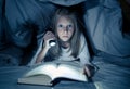 Scared little girl reading scary book under bed cover holding a flashlight late at night Royalty Free Stock Photo
