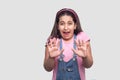 Scared sad brunette young girl in casual style, pink t-shirt and blue denim overalls standing with blocking hand and afraid face