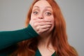 Scared young red hair woman covering her mouth with han