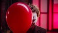 A scary clown peeking out from the red balloon and creepy smiling