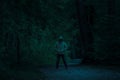 Scary dark male figure in a hoodie standing on the path in the forest with mysterious light coming behind him