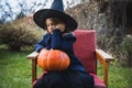 Scary little girl in witch costume, hat with big pumpkin celebrating halloween holiday. Sitting on armchair in coat with pumpkin. Royalty Free Stock Photo